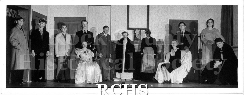 Black and white photograph of twelve cast members posed onstage. Most are standing, some are sitting. The set is an interior, furnished like a house.