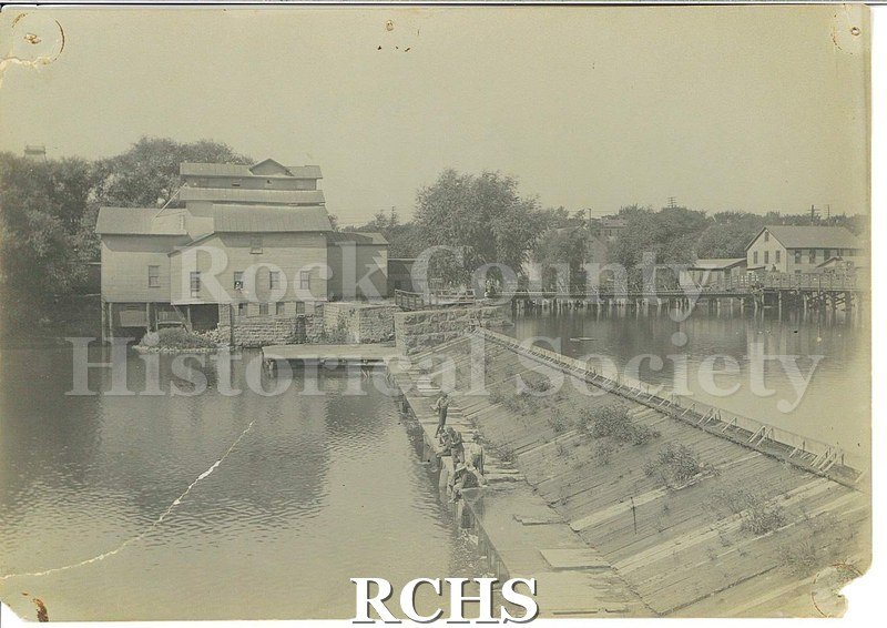 Black and white photograph of early Janesville dam with men fishing. Shows an unidentified building near the dam.