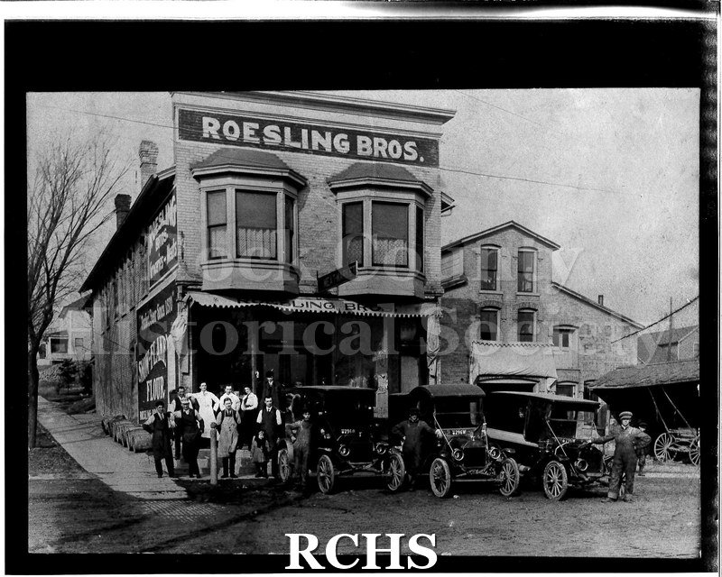 Black and white photograph of a group of 13 people stand outside on a street in front of a building, which has painted on top: "Roesling Bros." and another smaller.