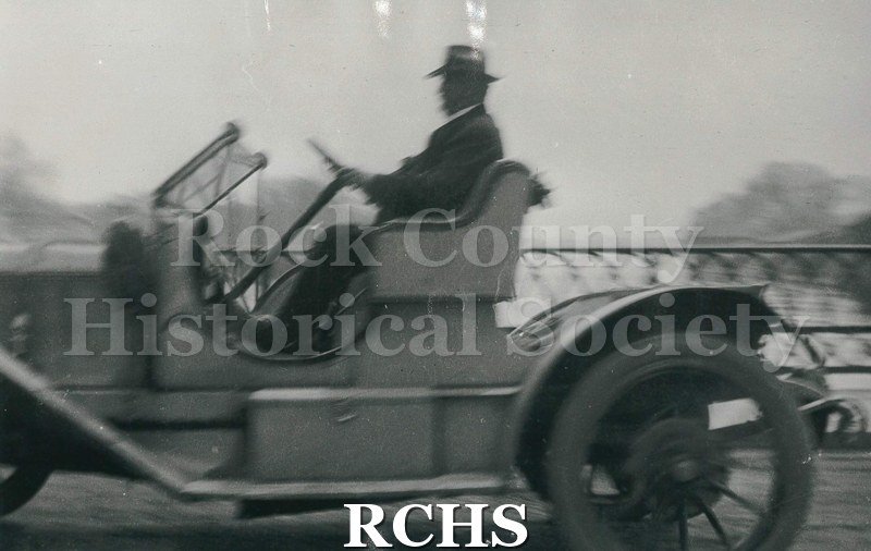 Blurry Black and white photograph of Dr. Hudley driving the first car in Janesville, circa 1907-1911. Make and model of the car is unknown.