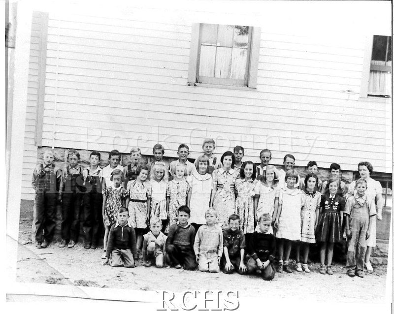 Black and white scan of a photo of a class of children and their teacher, standing outside their schoolhouse.