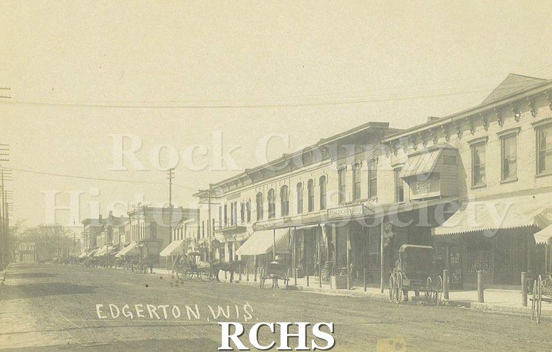 Postcard. Sepia-toned photographic image of downtown Edgerton, Wisconsin. There is a dirt road with numerous buggies outside local businesses.