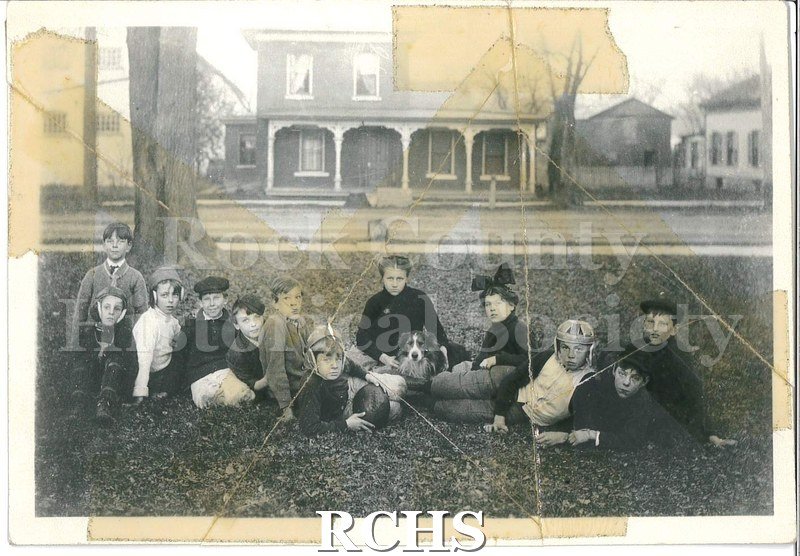 Black and white photograph of twelve children (ten boys, two girls) and a dog sitting on a lawn across the street from a house.