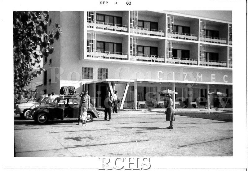 Black and white photograph of the outside of a hotel, with people standing around some parked cars.