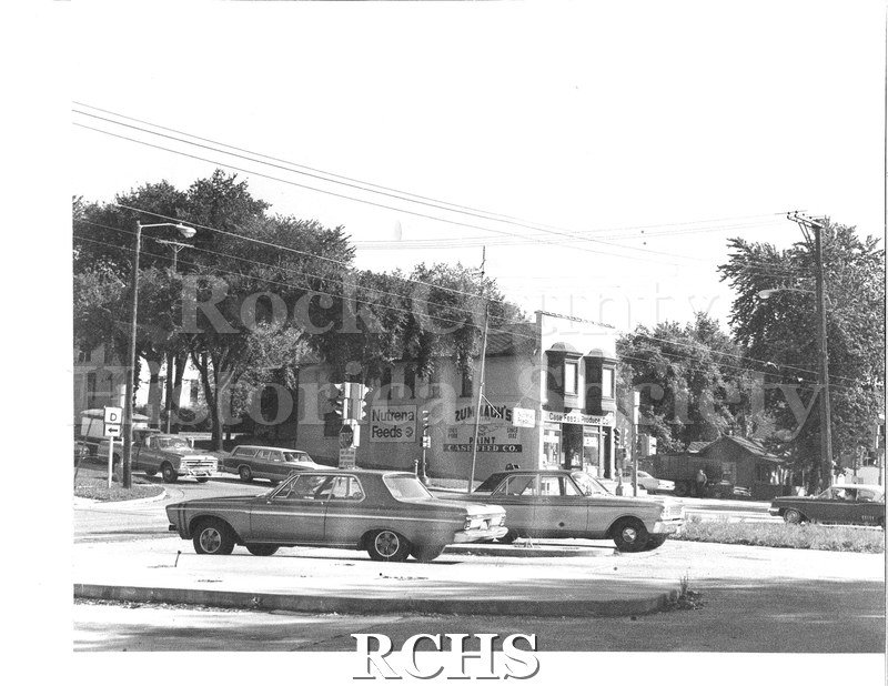 Black and white photograph of Case Feed & Produce Co. Shows a street corner with two cars parked in a lot, three cars stopped at a light. Case Feed & Produce Co.