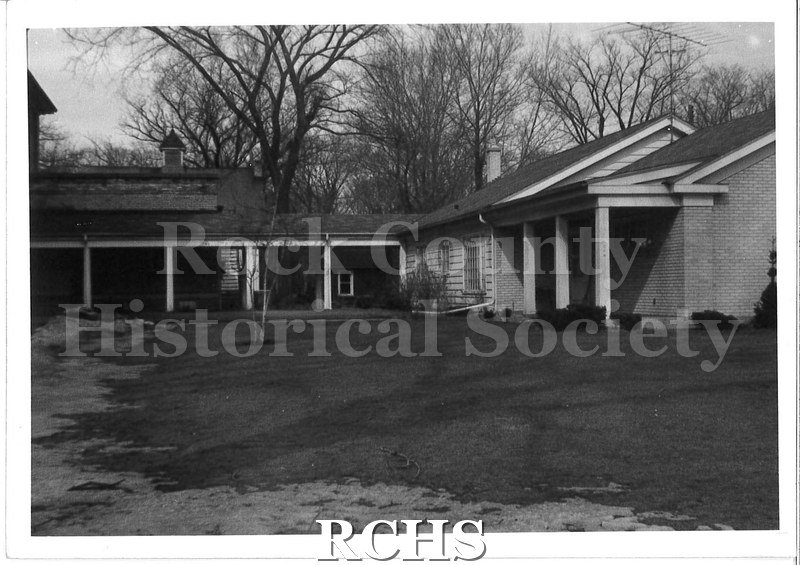 Black and white photograph of a long one story house with a breezeway on the left.