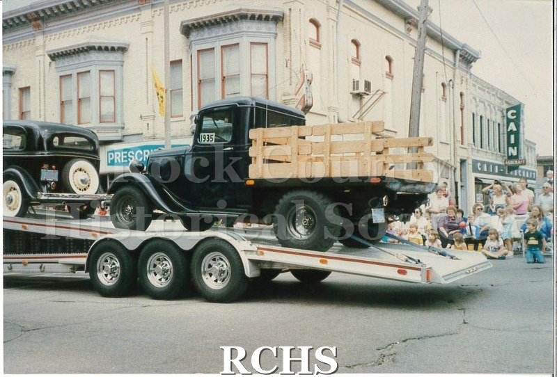 Colored photograph of the back end of a transport vehicle with an antique car on the front and an antique truck on the back.