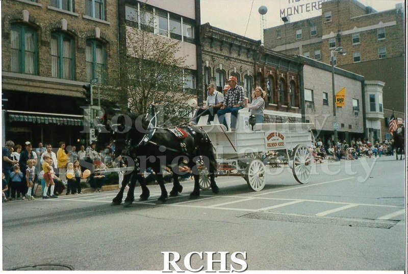 Colored photograph of two black horses pulling a white wagon along the parade route. There are several people riding in the wagon.