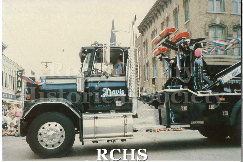Colored photograph of Davis Wrecker Service truck along the parade route. People are lining the route in front of Cain-Ashcroft.