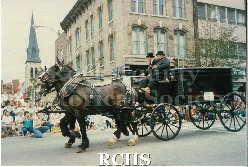 Colored photograph of two dark bay horses pulling a black hearse on four wheels. There are two men riding on the seat. There are people lining the parade route.