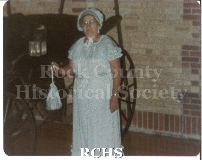 Colored photograph of an unidentified woman wearing a blue dress with a matching bonnet and purse, standing in front of a buggy parked inside a building.