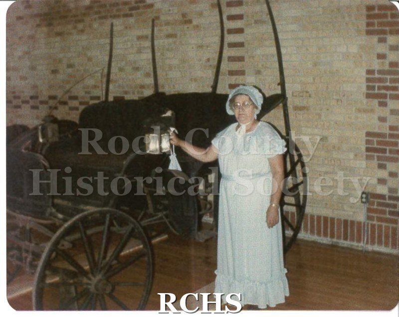 Colored photograph of an unidentified woman wearing a light blue dress, with a matching bonnet and purse standing in front of a buggy parked inside a building.