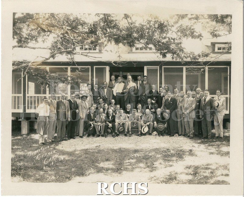 Black and white glossy photograph of the Rotary Club anniversary celebration showing several men standing and sitting in front of a building.