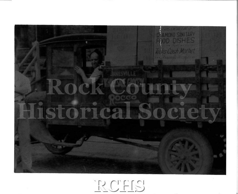 Black and white photograph of the cab of a truck. On the side of the truck is a sign: "Janesville / Wholesale Grocery / Company / Rocco".