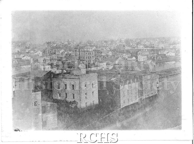 Black and white photograph of early Janesville. Shows many buildings along the river.
