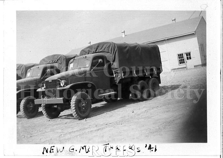 Black and white photograph of WWII Training Fort Knox, KY. New GMC cargo trucks parked in a row on a packed dirt courtyard.