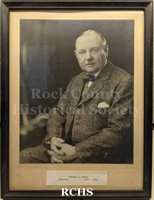 Back and white studio portrait of an older man in a suit, mounted behind glass in a black frame. The man is balding and has some grey or white hair, combed flat.