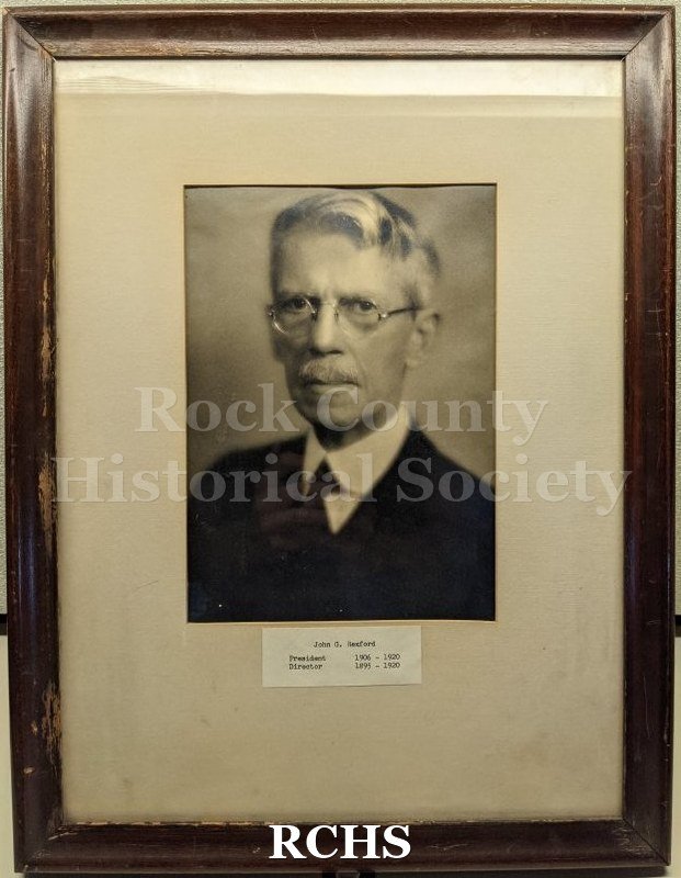 Black and white studio portrait bust of an old man in a suit, mounted behind glass in a brown wooden frame.