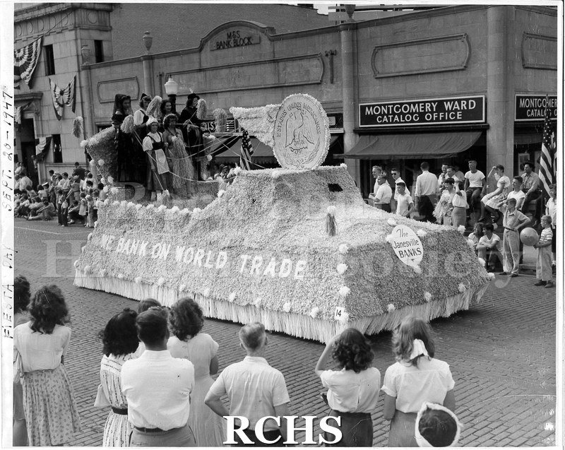 Black and white photograph of the Janesville Banks parade float, driving down a brick street as people watch from the sidewalks on Fiesta Days.