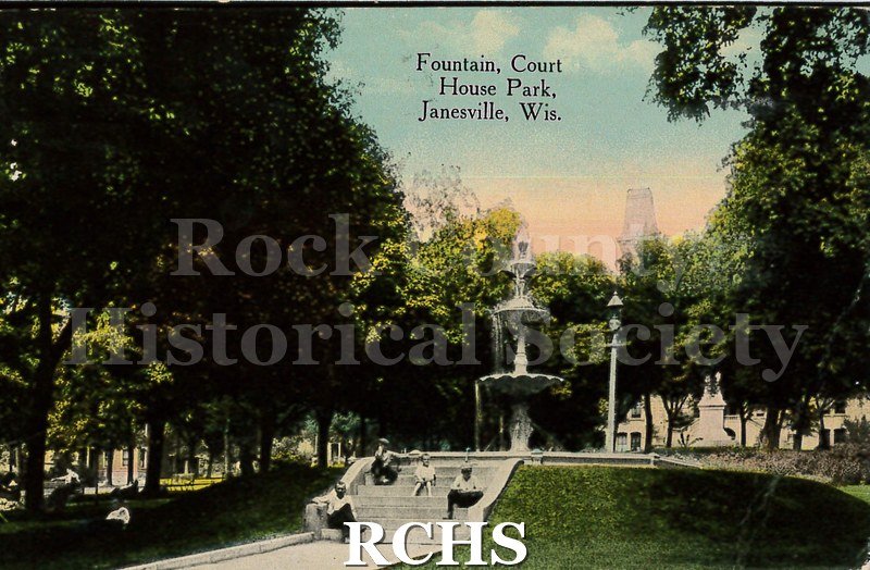 Postcard. Colorized image of the fountain in Court House Park in Janesville. Several men and boys are sitting on steps in front of the fountain.