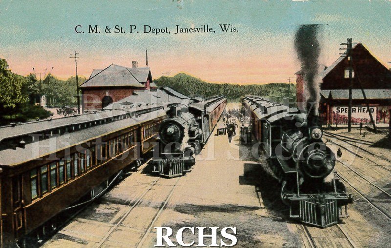Postcard. Colorized image of the Chicago, Milwaukee, & St. Paul Depot in Janesville. Image shows multiple steam locomotives with passenger cars attached.