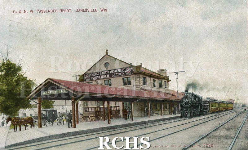 Postcard. Colorized image of the Chicago & Northwestern Railroad Depot in Janesville. Image shows a steam locomotive pulling up alongside the building.