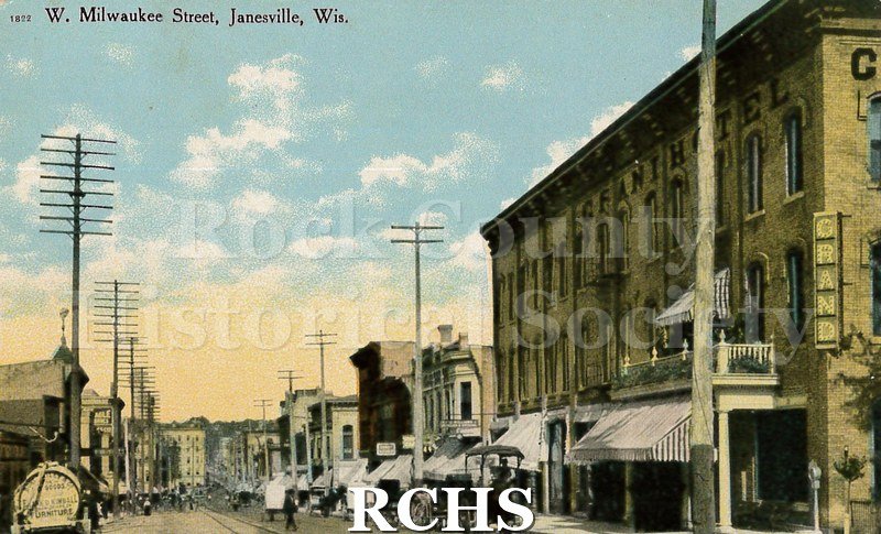 Postcard. View of W. Milwaukee Street in Janesville. Grand Hotel and other businesses visible. Street is full of carriages and pedestrians.