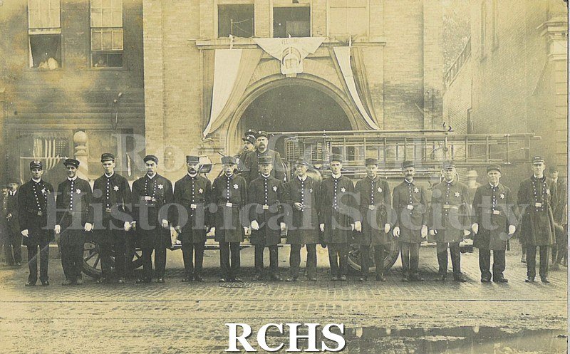 Black and white photograph of Janesville fire department members posed in front of a ladder truck with the fire station behind. C.