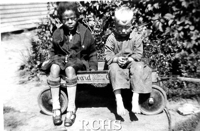 Black and white photograph of two children sitting in a wagon. Note on back says that the photo was probably taken by the Joyce family when they lived on Wall.