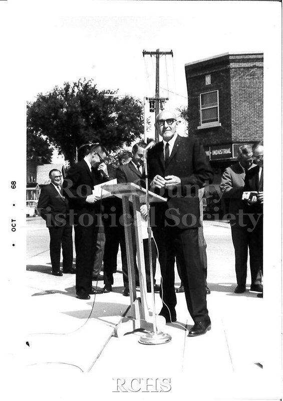 Black and white photograph of Joseph Lustig, the older man, standing at a podium outside. He is bald and is wearing glasses, a dark suit, and tie.