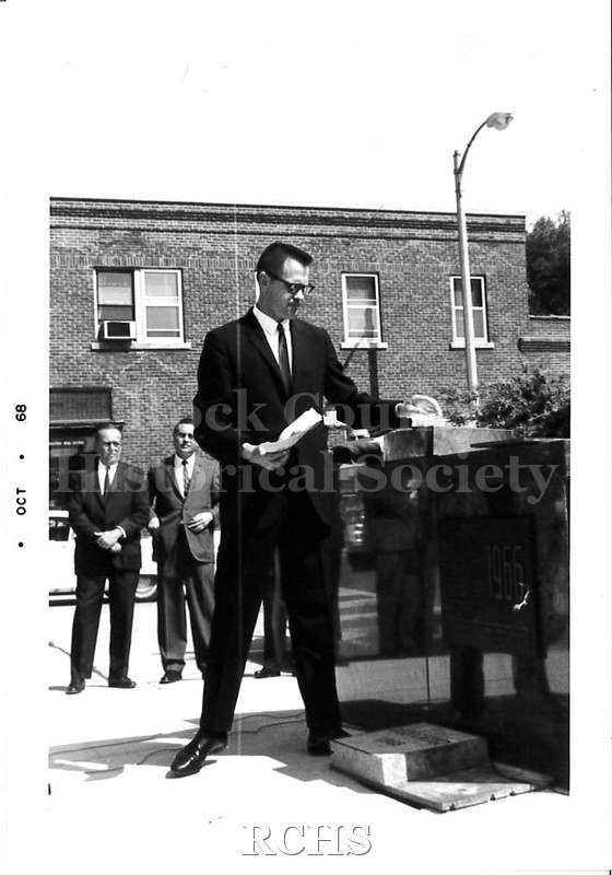 Black and white photograph of man in a suit standing next to a granite dedication slab outside.