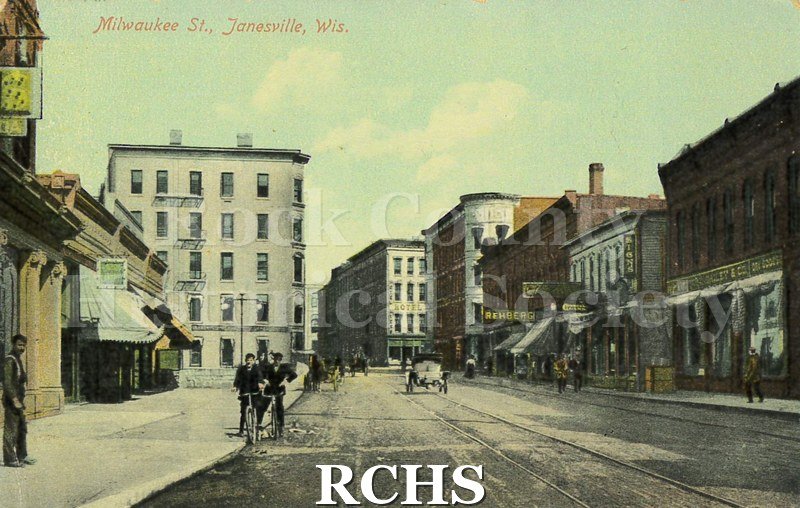 Postcard of Milwaukee Street. Storefronts, some people and a car are visible. Reverse has handwritten note in black ink addressed to "Mrs.