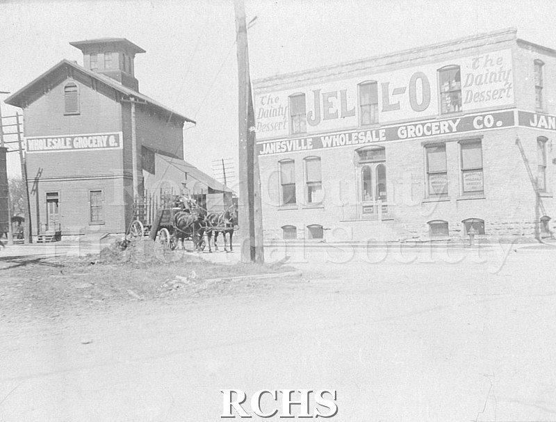 Postcard. Black and white photograph of a horse-drawn delivery carriage leaving the Janesville Wholesale Groecery Co., which was located at 202 N. High Street.