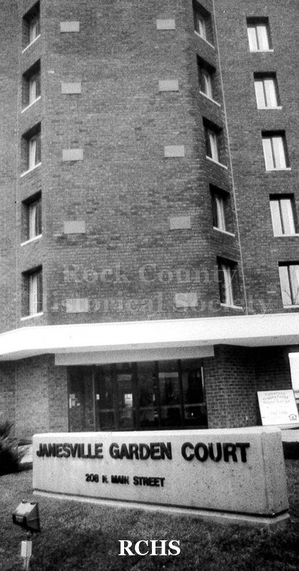 Black and white photograph of Janesville Garden Court apartment complex. In front of the building is a sign: "Janesville Garden Court 208 N. Main Street".