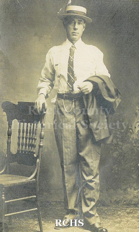 Black and white photograph of Rev. Wallace McGowan standing with his hand on a chair. He is holding his suit jacket over his arm.