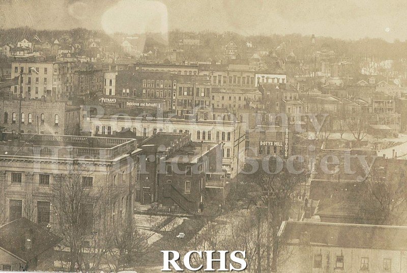 Black and white postcard of buildings and homes with bare trees and snow on the ground. Front left building appears to be old Post Office, looking east.