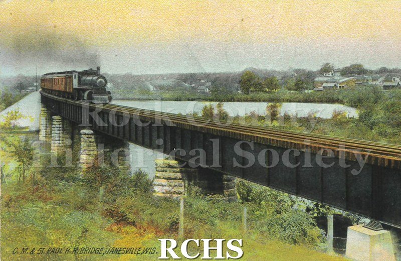 Postcard of C. and Saint Paul Bridge over river with hill with trees on left and building visible in background, especially on right. Train on bridge.