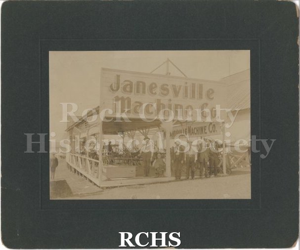 Sepia photo of Janesville Machine Company at an exhibition. Six men in hats and suits stand in front of an open-walled building.