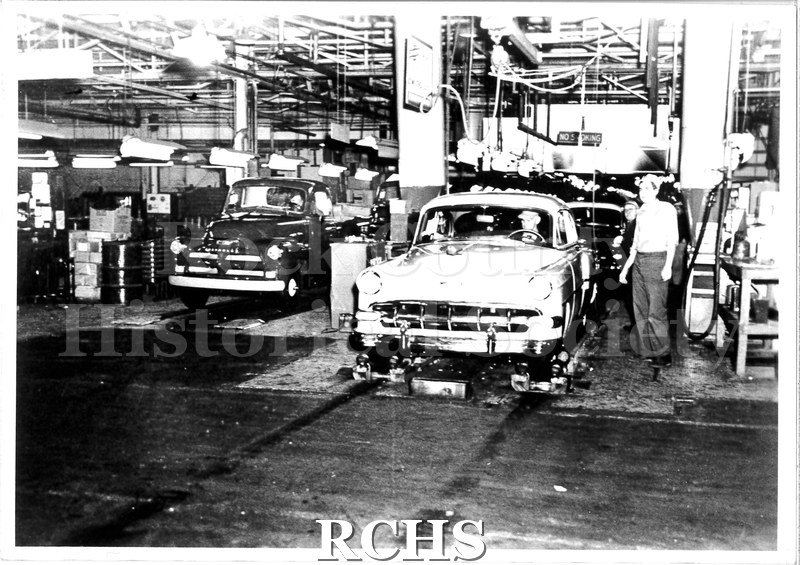 Black and white photograph of a car and a truck at the end of the Chevrolet assembly lines, 1954. The truck is a darker color, located on the left side of the photo.