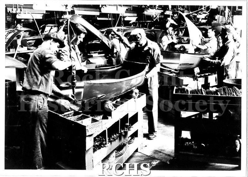 Black and white photograph of Chevrolet workers manufacturing car hoods. Three men work on each hood, some with power tools.