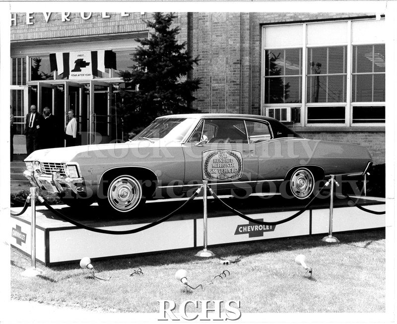 Black and white photograph of the 100 millionth GM vehicle on a podium outside the Janesville General Motors plant.