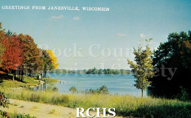 Postcard autumn lake scene. White text on front reads "Greetings from Janesville, Wisconsin". Reverse has no writing, postmark or stamp.
