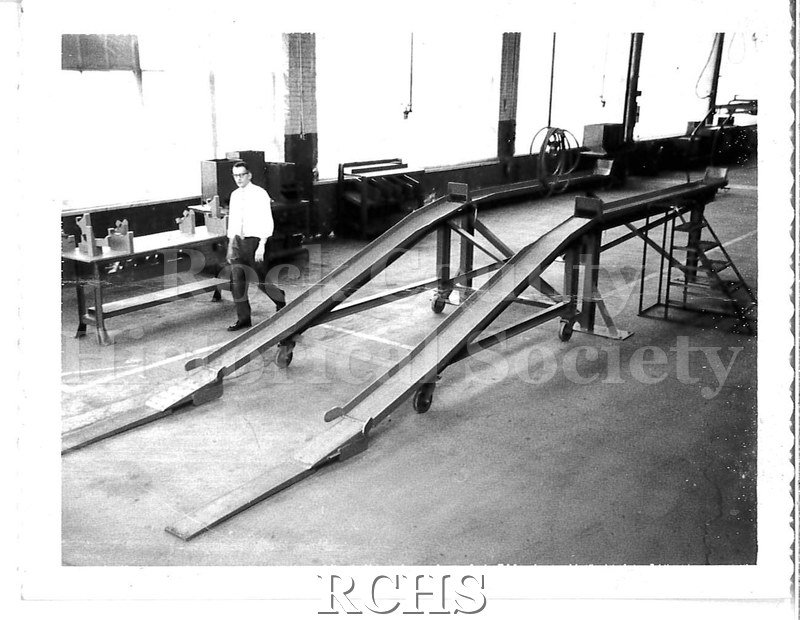 Black and white photograph of the General Motors factory floor. A man in a white shirt, tie, and slacks walks behind the ramp.