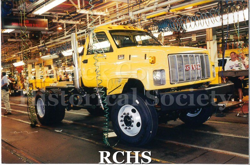Color photo of the Janesville GM plant assembly line for medium-duty trucks. A yellow truck (a GMC C8500) is coming down the line, not yet complete.