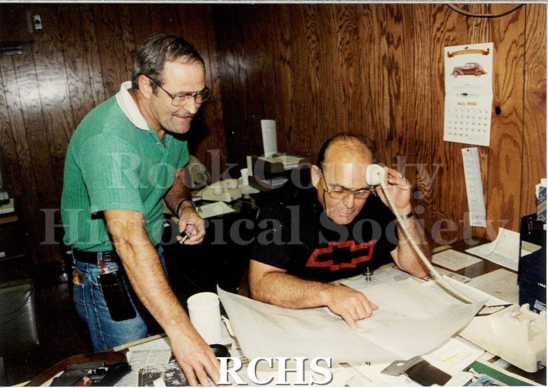 Color photo with flash of two men in the specs office at Janesville General Motors.
