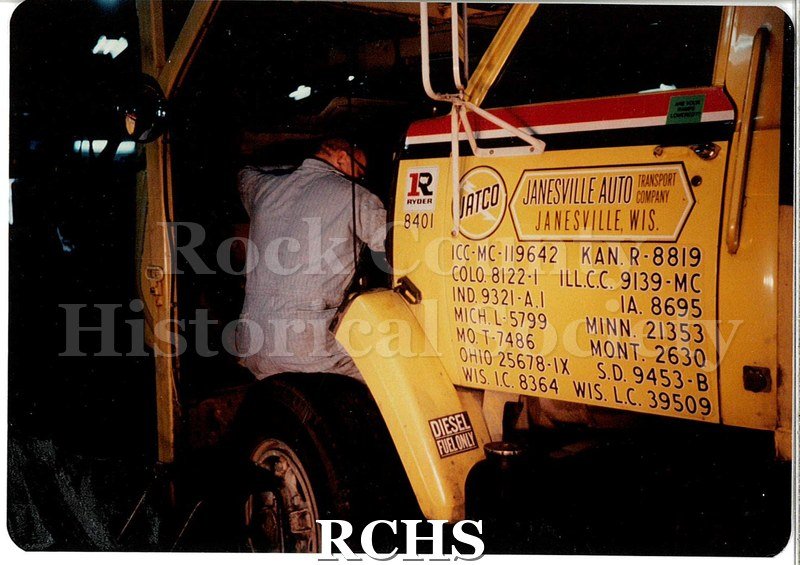 Color photo of a JATCO employee working under the hood of a yellow Ryder transport cab. He is wearing light blue coveralls and is up to his elbows in engine.