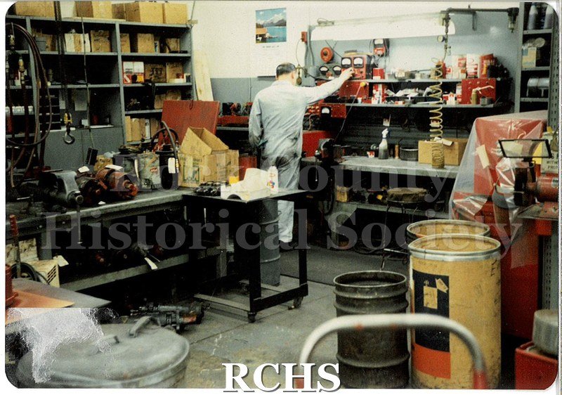 Color photo of a JATCO employee working in the engine shop. There are shelves against the left wall, filled with boxes of supplies.