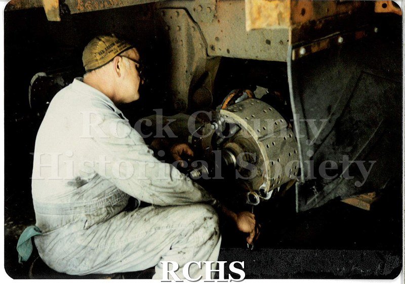 Color photo of a JATCO worker. He is wearing light blue coveralls, a brown skullcap, and glasses. He's seated and working on the brakes for a transport truck.