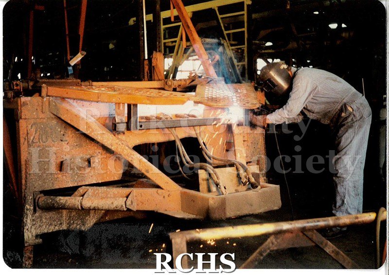 Color photo of a JATCO worker. He is welding something on a yellow transport trailer. Sparks fly, some spatter on the ground, and smoke curls into the air.
