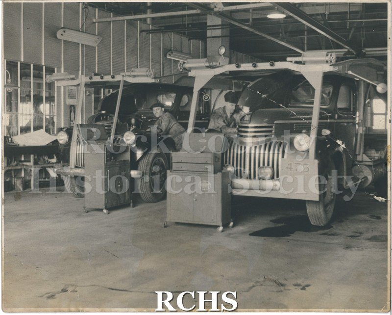 Black and white photograph of two men working on two transport trucks.